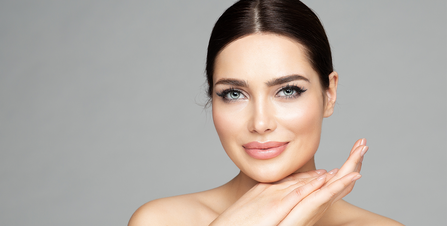 Elegant woman with flawless skin posing against gray background - Medical Spa & Wellness By Michelle NP Beautiful woman with smooth skin and striking blue eyes smiling softly with hands gently resting under her chin, set against a neutral gray backdrop - Medical Spa & Wellness By Michelle NP