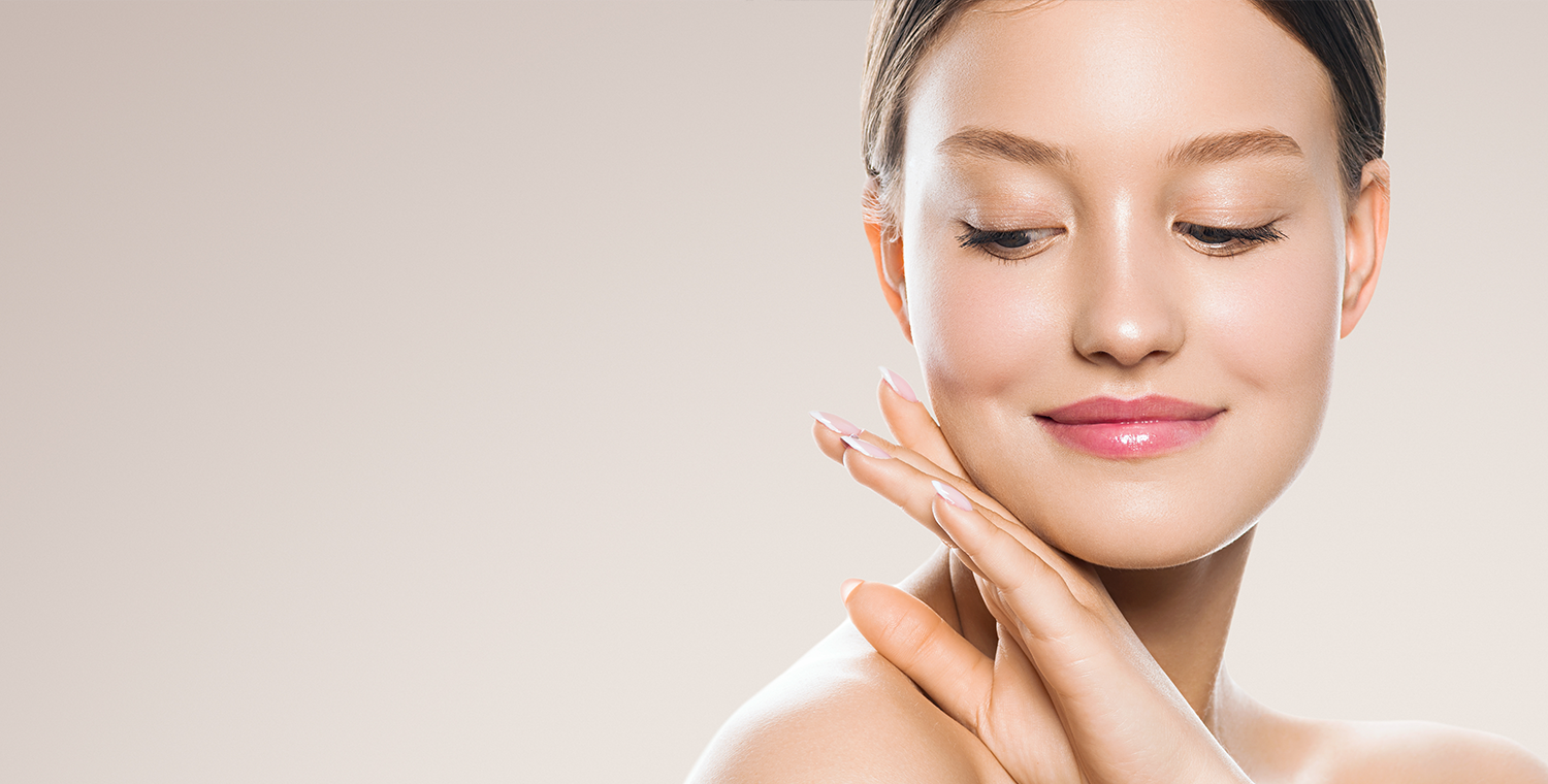 Graceful woman with smooth, glowing skin looking down - Medical Spa & Wellness By Michelle NP Young woman with flawless, radiant skin smiling softly while looking down and gently touching her chin with one hand, set against a neutral background - Medical Spa & Wellness By Michelle NP