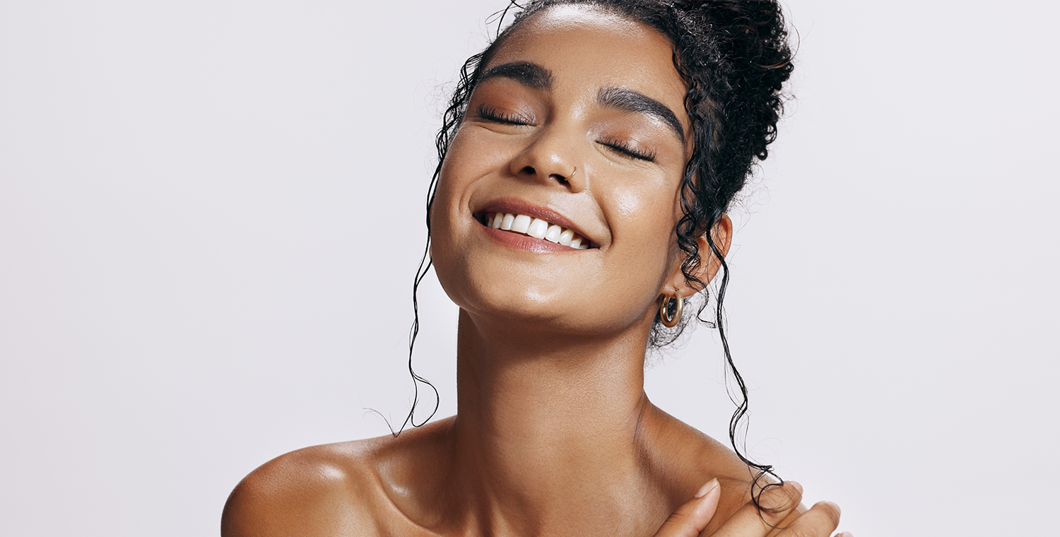 Joyful woman with glowing skin and closed eyes smiling - Medical Spa & Wellness By Michelle NP Radiant young woman with glowing skin and curly hair in a bun smiling with her eyes closed, exuding relaxation and confidence against a light background - Medical Spa & Wellness By Michelle NP